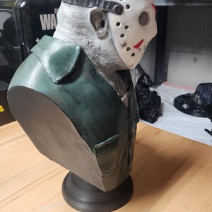 May include: A three-dimensional bust of a figure wearing a white hockey mask with red markings and a dark green collared jacket. The figure has a textured gray head and is mounted on a circular black base, viewed from a side profile angle.