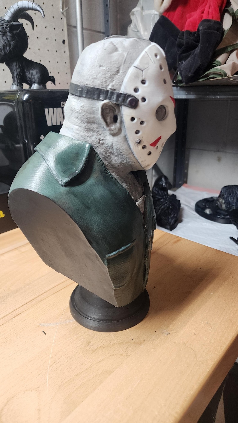 May include: A three-dimensional bust of a figure wearing a white hockey mask with red markings and a dark green collared jacket. The figure has a textured gray head and is mounted on a circular black base, viewed from a side profile angle.