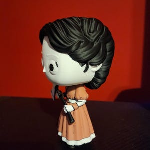 May include: A stylized vinyl figure with a large head and black hair, wearing a peach-colored dress with white trim. The figure has a white face with large black eyes and is holding a small black and white axe in its hand, viewed from the side.