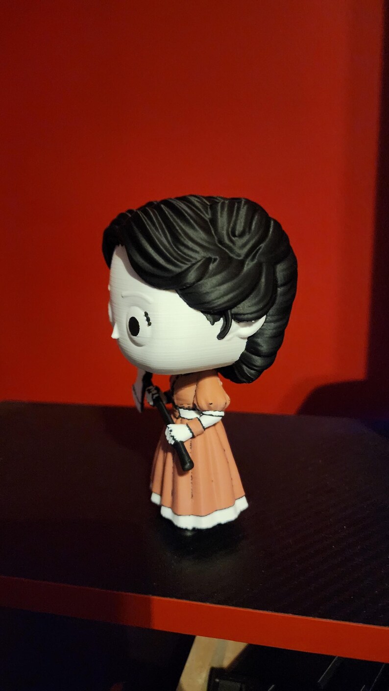 May include: A stylized vinyl figure with a large head and black hair, wearing a peach-colored dress with white trim. The figure has a white face with large black eyes and is holding a small black and white axe in its hand, viewed from the side.