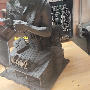 May include: A matte black resin sculpture depicting a character holding a chainsaw, standing on a cabin roof base with the text Evil Dead 2. The figure features a detailed shirt, a book with a face on the cover, and a chimney on the cabin structure.