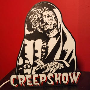 May include: A black and white wall sign featuring a graphic illustration of a skeletal figure in a hooded robe with the text CREEPSHOW printed in bold, block letters at the bottom. The sign has a white power cord extending from the left side.