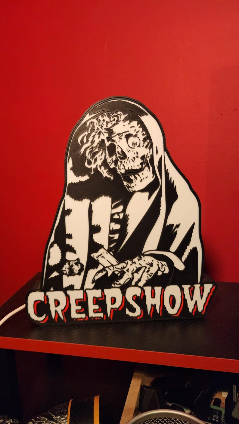 May include: A black and white wall sign featuring a graphic illustration of a skeletal figure in a hooded robe with the text CREEPSHOW printed in bold, block letters at the bottom. The sign has a white power cord extending from the left side.