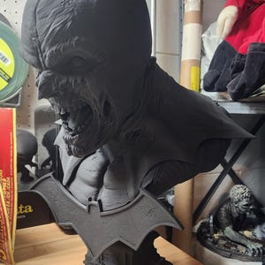May include: A matte black 3D printed bust of a zombie-like Batman character with a snarling expression, pointed ears, and a bat-shaped emblem on the base. The base features small skull details and a tombstone design, rendered in a solid dark finish.
