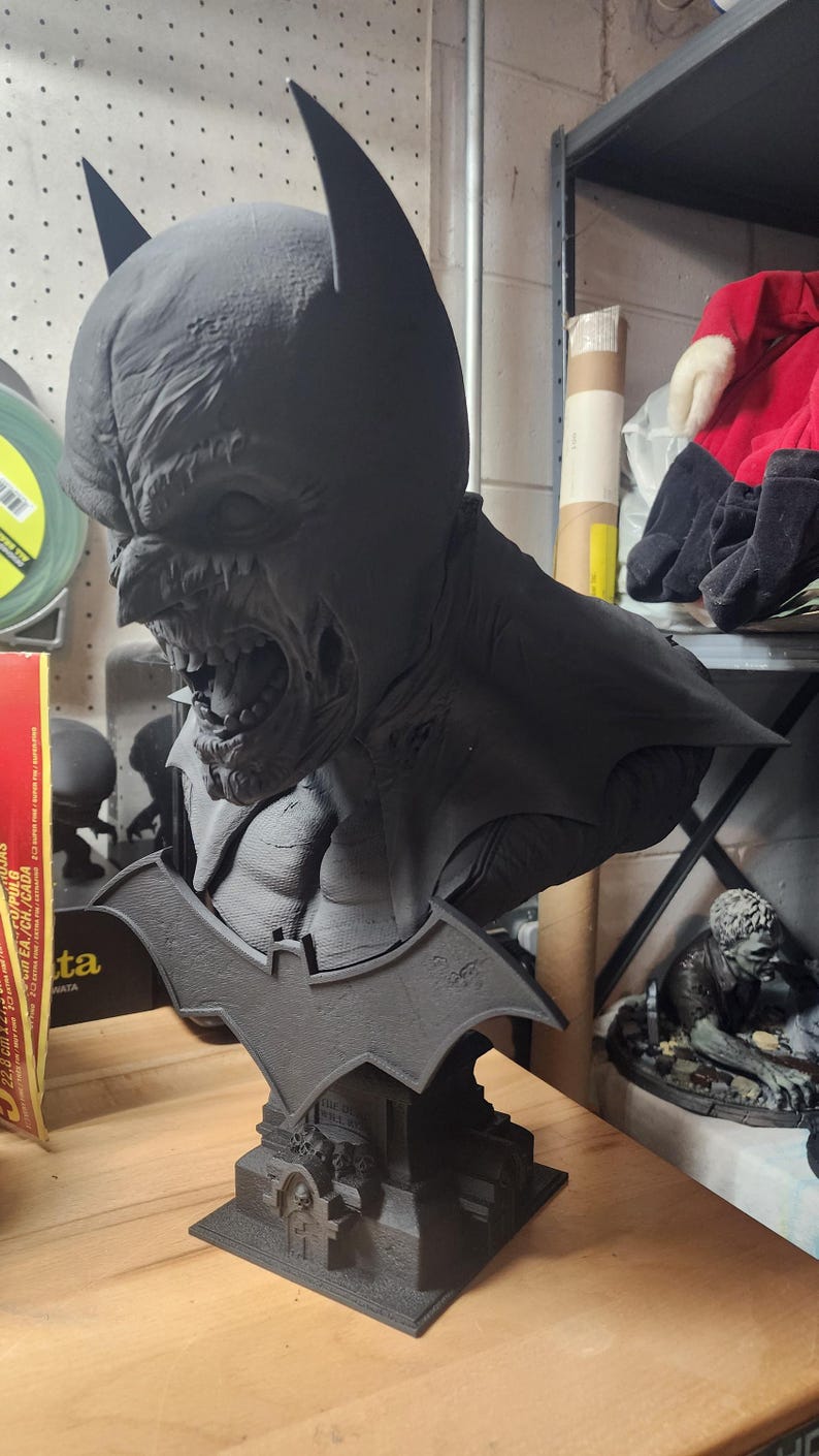 May include: A matte black 3D printed bust of a zombie-like Batman character with a snarling expression, pointed ears, and a bat-shaped emblem on the base. The base features small skull details and a tombstone design, rendered in a solid dark finish.
