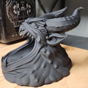 May include: A matte black 3D printed sculpture of a horned creature with an open mouth, featuring sharp teeth, pointed ears, and a textured base. The creature has a wide, hollowed-out jaw area and prominent curved horns extending from its head.