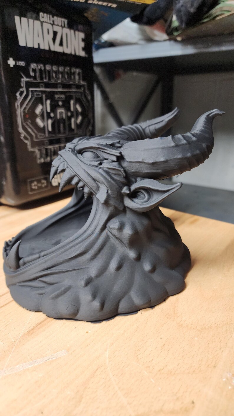 May include: A matte black 3D printed sculpture of a horned creature with an open mouth, featuring sharp teeth, pointed ears, and a textured base. The creature has a wide, hollowed-out jaw area and prominent curved horns extending from its head.