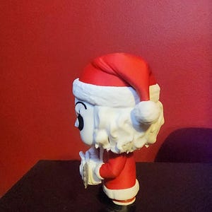May include: A stylized vinyl figure of a character with white hair and a pale face, wearing a red and white Santa Claus hat and a matching red suit with white trim. The figure is shown in profile, holding a small white object in its hands.