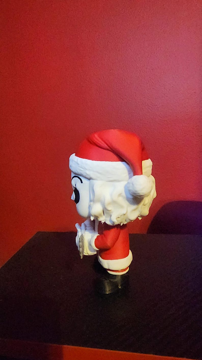 May include: A stylized vinyl figure of a character with white hair and a pale face, wearing a red and white Santa Claus hat and a matching red suit with white trim. The figure is shown in profile, holding a small white object in its hands.