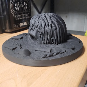 May include: A matte black 3D printed sculpture featuring a human head partially submerged in a textured base. The base includes sculpted details of wooden planks, a chain, and a smooth stone. The head has long, flowing hair and a neutral expression.