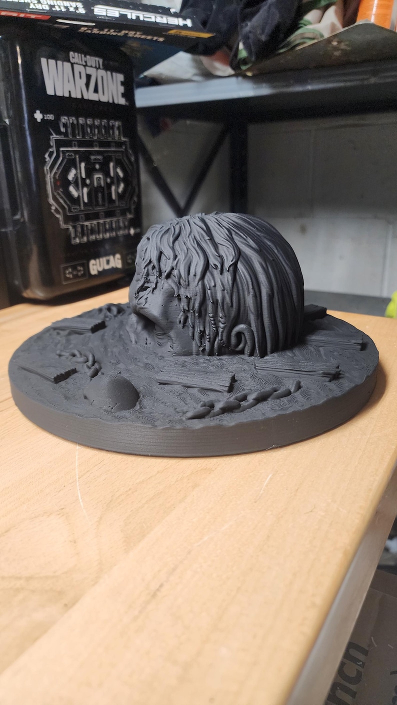 May include: A matte black 3D printed sculpture featuring a human head partially submerged in a textured base. The base includes sculpted details of wooden planks, a chain, and a smooth stone. The head has long, flowing hair and a neutral expression.