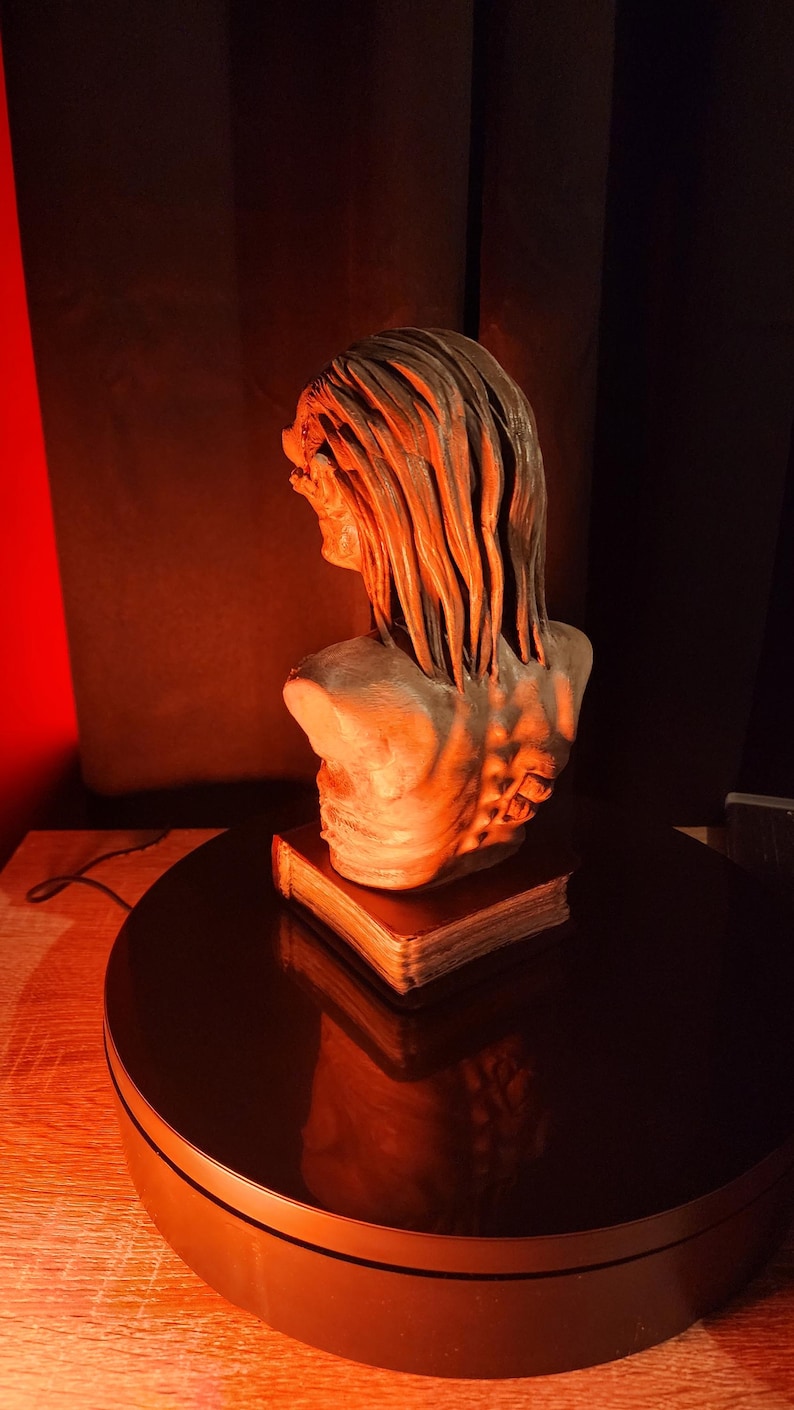 May include: A grey-toned bust sculpture of a figure with long, textured hair and a visible spinal column, mounted on top of a rectangular book. The sculpture features intricate details on the back and shoulders, presented in a monochromatic finish.