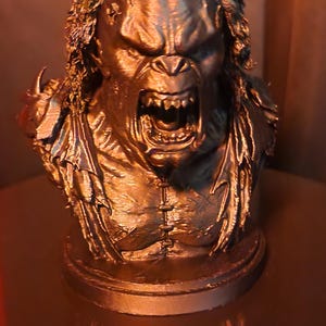 May include: A bronze-coloured bust of a creature with long, textured hair, a prominent brow, and an open mouth revealing sharp teeth. The figure features a muscular chest with visible stitching details and jagged, torn clothing elements on the shoulders.