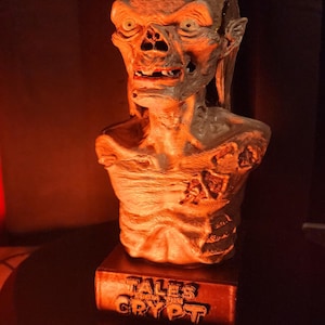 May include: A sculpted bust of the Crypt Keeper character featuring a weathered, skeletal appearance with sunken eyes and exposed teeth. The figure is mounted on a rectangular base with the embossed text TALES FROM THE CRYPT in a dark finish.