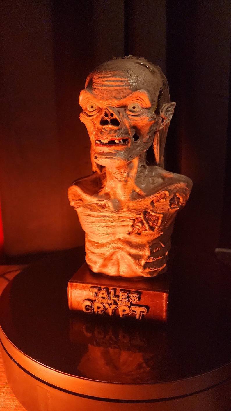 May include: A sculpted bust of the Crypt Keeper character featuring a weathered, skeletal appearance with sunken eyes and exposed teeth. The figure is mounted on a rectangular base with the embossed text TALES FROM THE CRYPT in a dark finish.