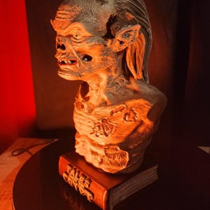 May include: A sculpted bust of a creature with textured, weathered skin and long hair, mounted on a base shaped like a book with the text TALES FROM THE CRYPT embossed on the spine. The figure features deep-set eyes, a prominent brow, and exposed teeth.
