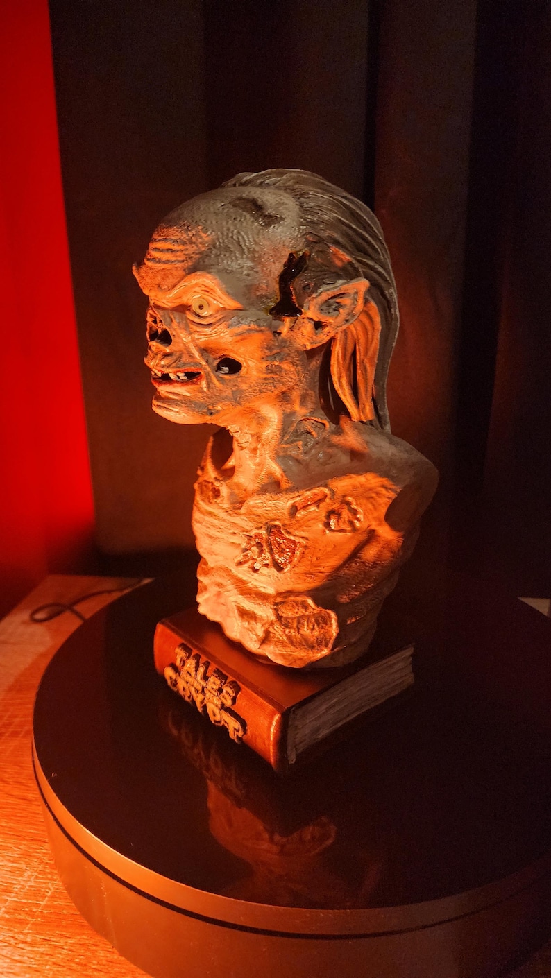 May include: A sculpted bust of a creature with textured, weathered skin and long hair, mounted on a base shaped like a book with the text TALES FROM THE CRYPT embossed on the spine. The figure features deep-set eyes, a prominent brow, and exposed teeth.