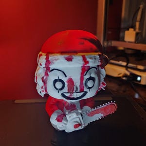 May include: A stylized vinyl figure of a character wearing a red Santa hat and red clothing, featuring a white face with black eye markings and a wide grin. The figure is covered in red paint splatters and holds a white chainsaw with red paint.