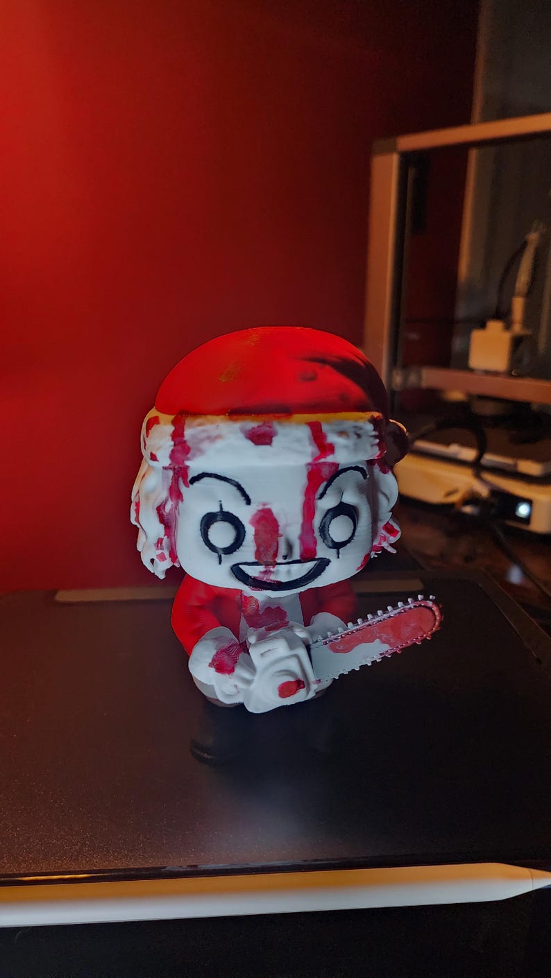 May include: A stylized vinyl figure of a character wearing a red Santa hat and red clothing, featuring a white face with black eye markings and a wide grin. The figure is covered in red paint splatters and holds a white chainsaw with red paint.