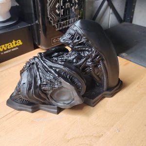 May include: A black, 3D-printed Xenomorph figure from the Alien franchise, featuring a smooth, elongated head, intricate ribbed tubing, and a segmented, biomechanical body structure. The figure is displayed in a curled, resting position on a base.