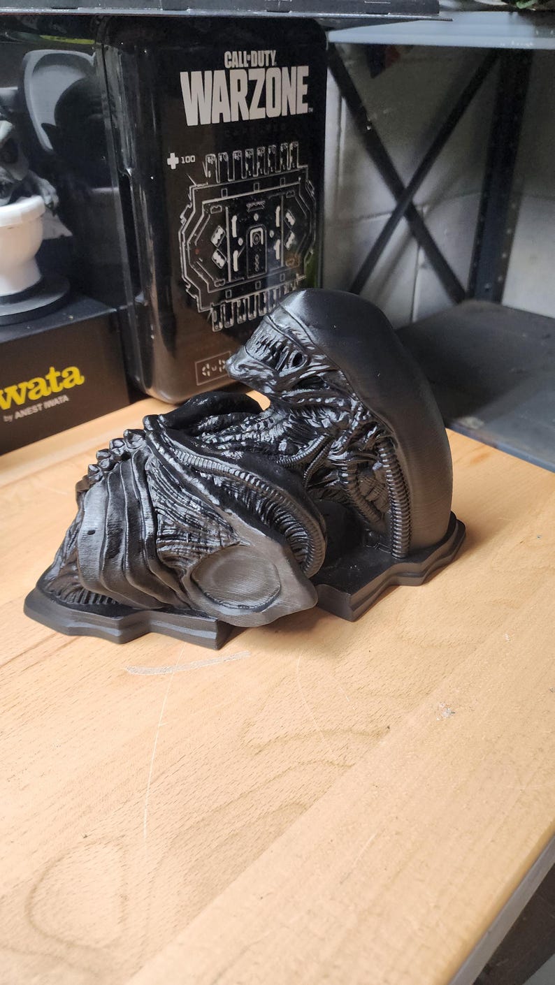 May include: A black, 3D-printed Xenomorph figure from the Alien franchise, featuring a smooth, elongated head, intricate ribbed tubing, and a segmented, biomechanical body structure. The figure is displayed in a curled, resting position on a base.