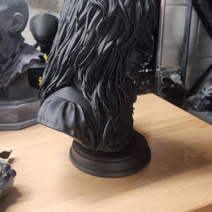 May include: A matte black 3D printed bust of a figure with long, wavy, textured hair and a detailed garment. The sculpture is mounted on a circular base and features intricate hair strands and fabric folds, captured from a side profile angle.