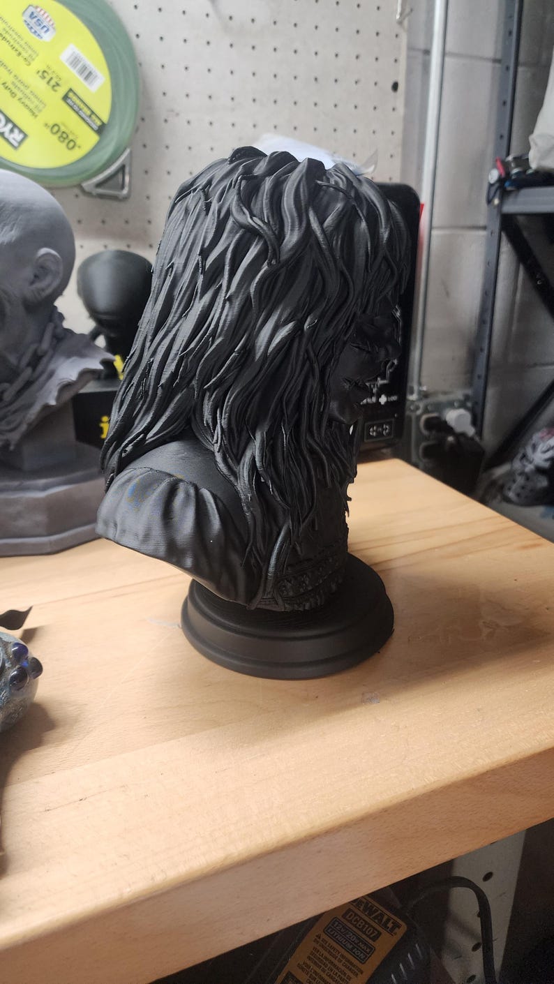 May include: A matte black 3D printed bust of a figure with long, wavy, textured hair and a detailed garment. The sculpture is mounted on a circular base and features intricate hair strands and fabric folds, captured from a side profile angle.