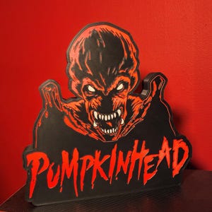 May include: A black and red wall sign featuring a graphic illustration of a creature with sharp teeth and glowing eyes, positioned above the text PUMPKINHEAD in a stylized, dripping red font. The sign has a raised, layered design.