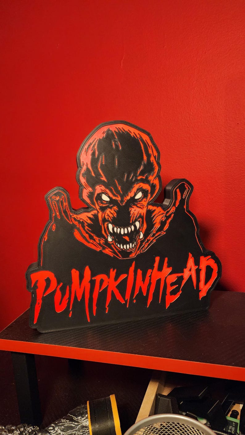 May include: A black and red wall sign featuring a graphic illustration of a creature with sharp teeth and glowing eyes, positioned above the text PUMPKINHEAD in a stylized, dripping red font. The sign has a raised, layered design.