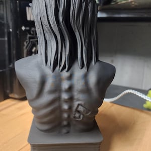 May include: A matte black 3D printed bust of a figure with long, textured hair and a visible spine. The figure has a prominent number 5 embossed on the right shoulder blade area. The bust is mounted on a rectangular base with horizontal ridges.