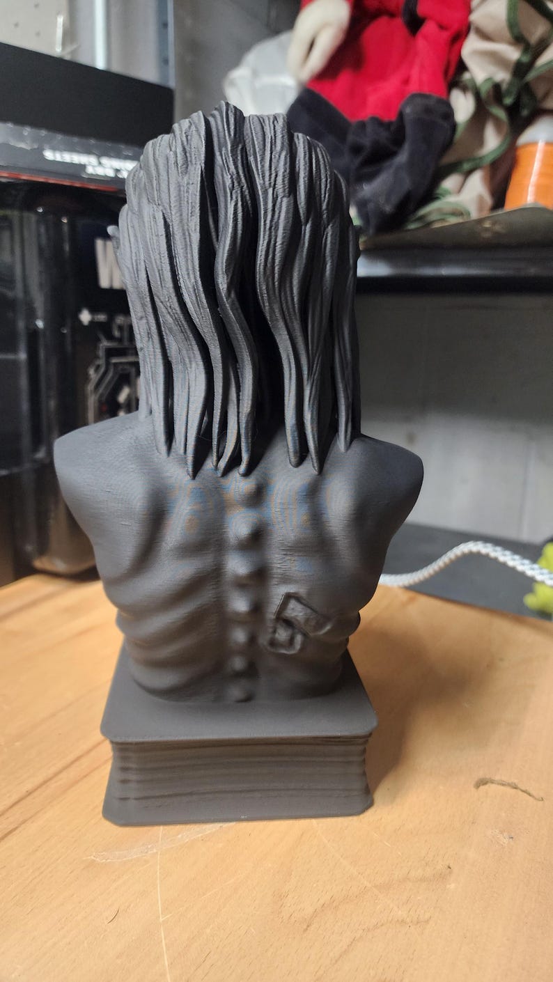 May include: A matte black 3D printed bust of a figure with long, textured hair and a visible spine. The figure has a prominent number 5 embossed on the right shoulder blade area. The bust is mounted on a rectangular base with horizontal ridges.