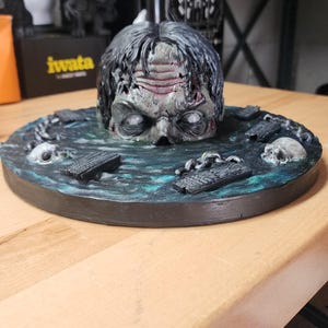 May include: A sculpted figure of a zombie head emerging from a dark, textured pool of water. The base features scattered debris, including small skulls and rectangular planks, all painted in shades of deep blue, black, and grey with white eyes.