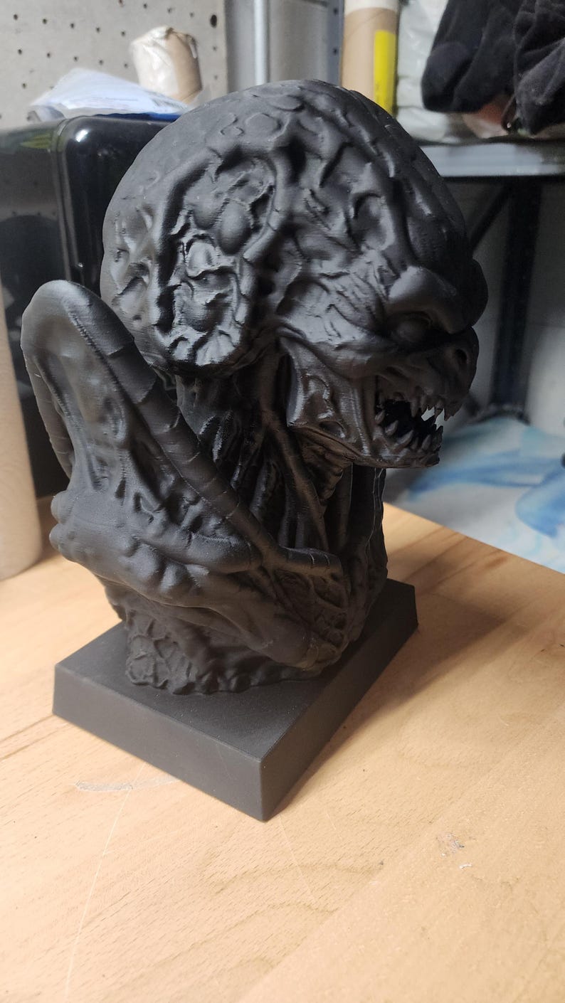 Puede incluir: Un busto negro mate impreso en 3D de una criatura con un cr&aacute;neo texturizado similar a un cerebro, dientes afilados y una mand&iacute;bula prominente. La figura presenta crestas y pliegues org&aacute;nicos complejos en toda su superficie sobre una base.
