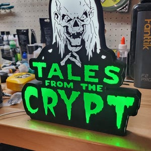 May include: A black and green illuminated sign featuring the white graphic of a skeletal figure with long hair above the text "TALES FROM THE CRYPT" in a dripping font. The sign is constructed with a black border and bright green light.
