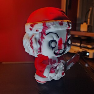 May include: A stylized vinyl figure of a character wearing a red and white Santa Claus suit, featuring a white face with black eye markings and a wide grin. The figure is covered in red blood-like splatters and holds a small grey chainsaw.