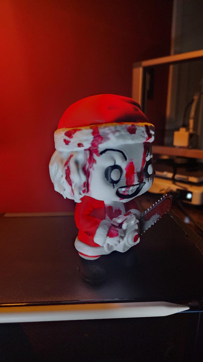 May include: A stylized vinyl figure of a character wearing a red and white Santa Claus suit, featuring a white face with black eye markings and a wide grin. The figure is covered in red blood-like splatters and holds a small grey chainsaw.
