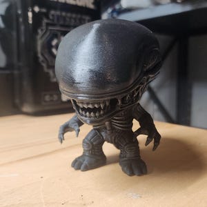 May include: A black, stylized figurine of a Xenomorph creature with an elongated, smooth head, visible teeth, and clawed hands. The figure has a textured, dark finish and stands in a crouched position with a detailed, segmented torso and limbs.