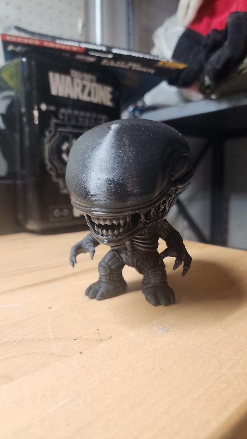 May include: A black, stylized figurine of a Xenomorph creature with an elongated, smooth head, visible teeth, and clawed hands. The figure has a textured, dark finish and stands in a crouched position with a detailed, segmented torso and limbs.