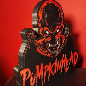 May include: A three-dimensional wall sign featuring the Pumpkinhead character in black and red. The design displays a menacing creature with sharp teeth and glowing white eyes, with the word PUMPKINHEAD written in a jagged, red font below.