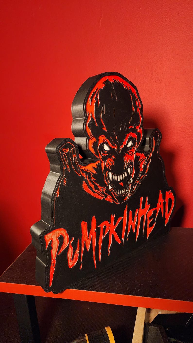 May include: A three-dimensional wall sign featuring the Pumpkinhead character in black and red. The design displays a menacing creature with sharp teeth and glowing white eyes, with the word PUMPKINHEAD written in a jagged, red font below.