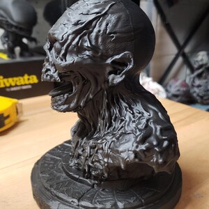 Op de afbeelding: Een matzwart 3D-geprint borstbeeld van een wezen met een textuur van aders, een open mond en een prominente kaaklijn. Het figuur is gemonteerd op een ronde voet met een radiaal, spinnenwebachtig patroon in het bovenoppervlak gegraveerd.