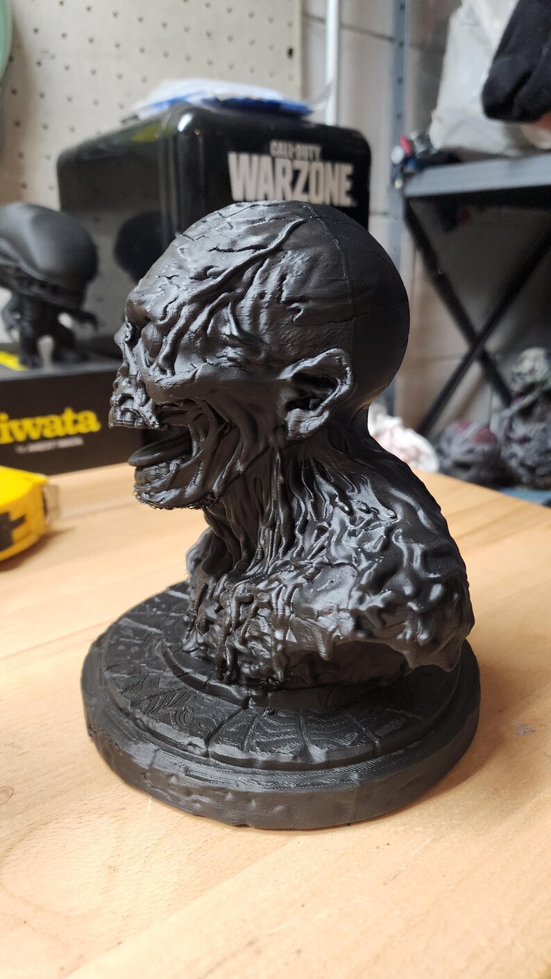 Op de afbeelding: Een matzwart 3D-geprint borstbeeld van een wezen met een textuur van aders, een open mond en een prominente kaaklijn. Het figuur is gemonteerd op een ronde voet met een radiaal, spinnenwebachtig patroon in het bovenoppervlak gegraveerd.