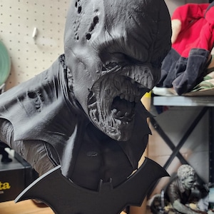 May include: A matte black 3D printed bust of a zombie Batman character. The figure features a damaged cowl with holes, exposed teeth, and a decaying face. A bat-shaped emblem is attached to the front of the base, which includes small sculpted tombstones.