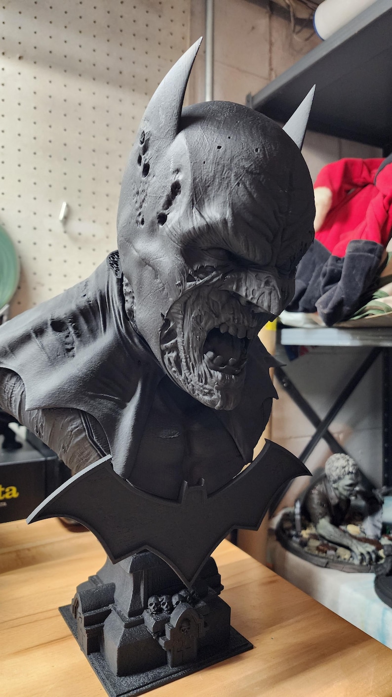 May include: A matte black 3D printed bust of a zombie Batman character. The figure features a damaged cowl with holes, exposed teeth, and a decaying face. A bat-shaped emblem is attached to the front of the base, which includes small sculpted tombstones.