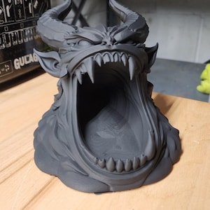 May include: A matte dark gray 3D printed sculpture of a horned demon head with a wide-open mouth, featuring sharp teeth and textured details. The text FILAMENT POOP EATING DEMON is visible at the top, with dimensions of 5.03 in height, 5.61 in width, and 6.65 in depth.