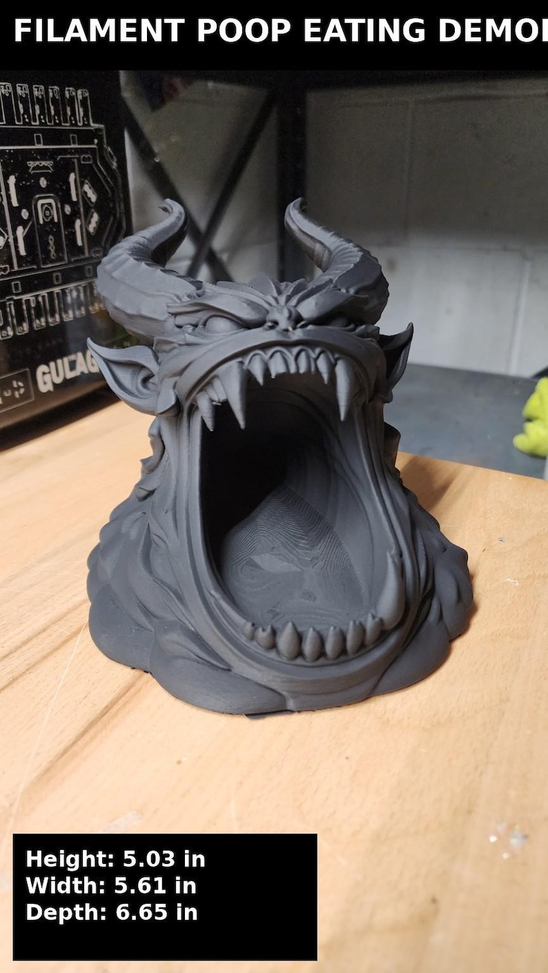 May include: A matte dark gray 3D printed sculpture of a horned demon head with a wide-open mouth, featuring sharp teeth and textured details. The text FILAMENT POOP EATING DEMON is visible at the top, with dimensions of 5.03 in height, 5.61 in width, and 6.65 in depth.