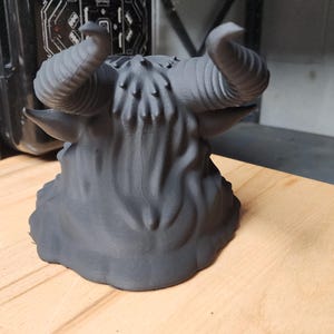 May include: A matte black 3D printed sculpture of a horned creature head, featuring prominent curved horns, textured fur-like details on the crown, and pointed ears. The object is shown from a rear-facing perspective, displaying a smooth base.