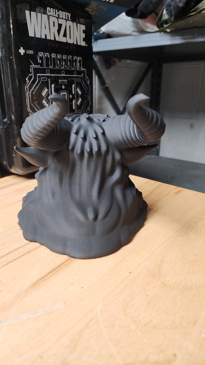May include: A matte black 3D printed sculpture of a horned creature head, featuring prominent curved horns, textured fur-like details on the crown, and pointed ears. The object is shown from a rear-facing perspective, displaying a smooth base.
