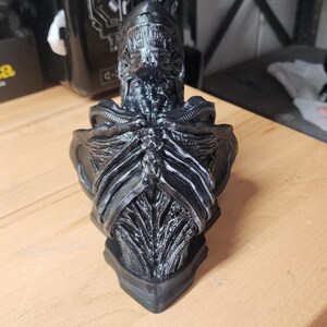 May include: A black, highly detailed bust of a Xenomorph creature from the Alien franchise. The sculpture features a smooth, elongated head, exposed rib-like structures on the chest, and intricate, biomechanical textures across the entire surface.