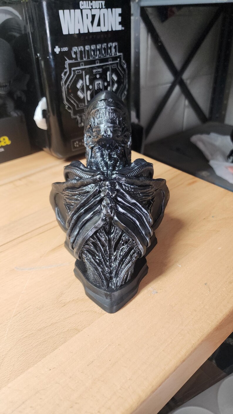 May include: A black, highly detailed bust of a Xenomorph creature from the Alien franchise. The sculpture features a smooth, elongated head, exposed rib-like structures on the chest, and intricate, biomechanical textures across the entire surface.